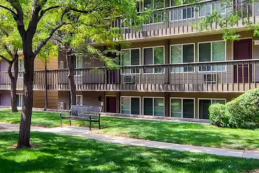 Martin Acres neighborhood in South Boulder, Colorado — three-story garden-style condo building with wood-clad balconies, mature tree canopy, and a bench on a landscaped walkway