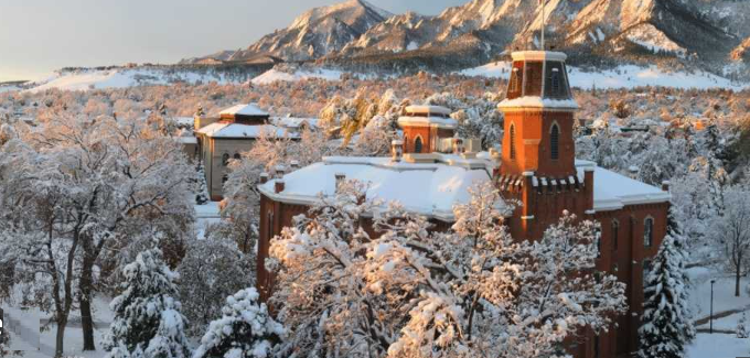 University of Colorado Boulder campus in winter — Old Main's red sandstone turrets and snow-covered Flatirons rising behind, viewed from the University Hill neighborhood